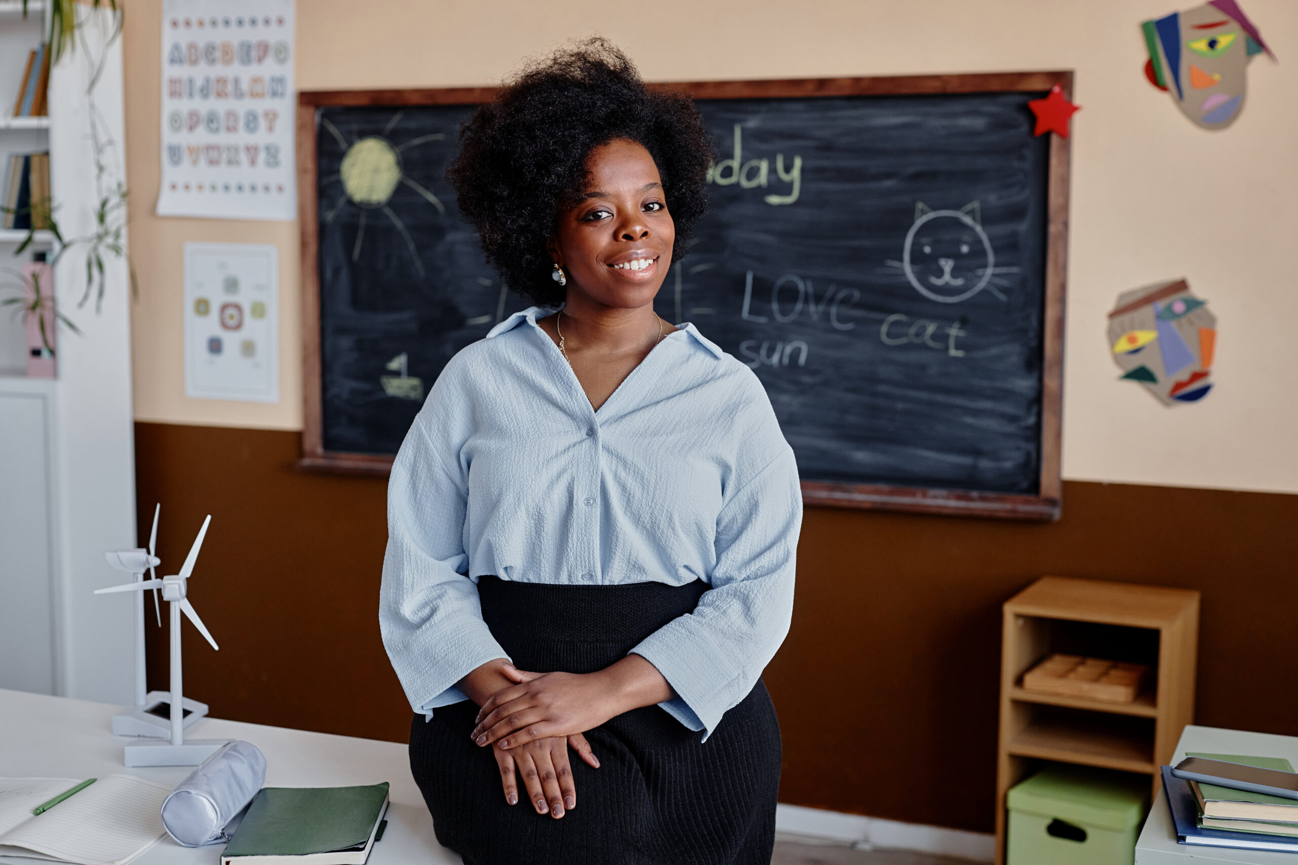 Portrait of Smiling Primary School Teacher in Classroom Medium long shot portrait of happy Black woman working as primary school teacher sitting on desk looking at camera in classroom, copy space
